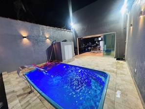 A swimming pool with a tiled deck, a covered patio area, and a view into a kitchen. - Perfect com piscina e 3-bedroom house in Peruíbe for a relaxing getaway (Peruíbe)