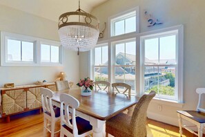 A dining area with a wooden table, white chairs, a chandelier, and large windows with a view of buildings. - Stunning Intracoastal Views + Sunset Porch | Easy Beach Access | 5BR | Sleeps 12 (Wrightsville Beach)