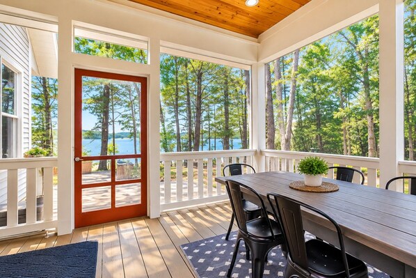 A covered deck with a table and chairs, overlooking a lake with trees.