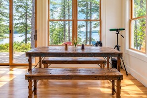 Una mesa de comedor de madera con bancos en una habitación con ventanas amplias que dan a un lago y árboles.