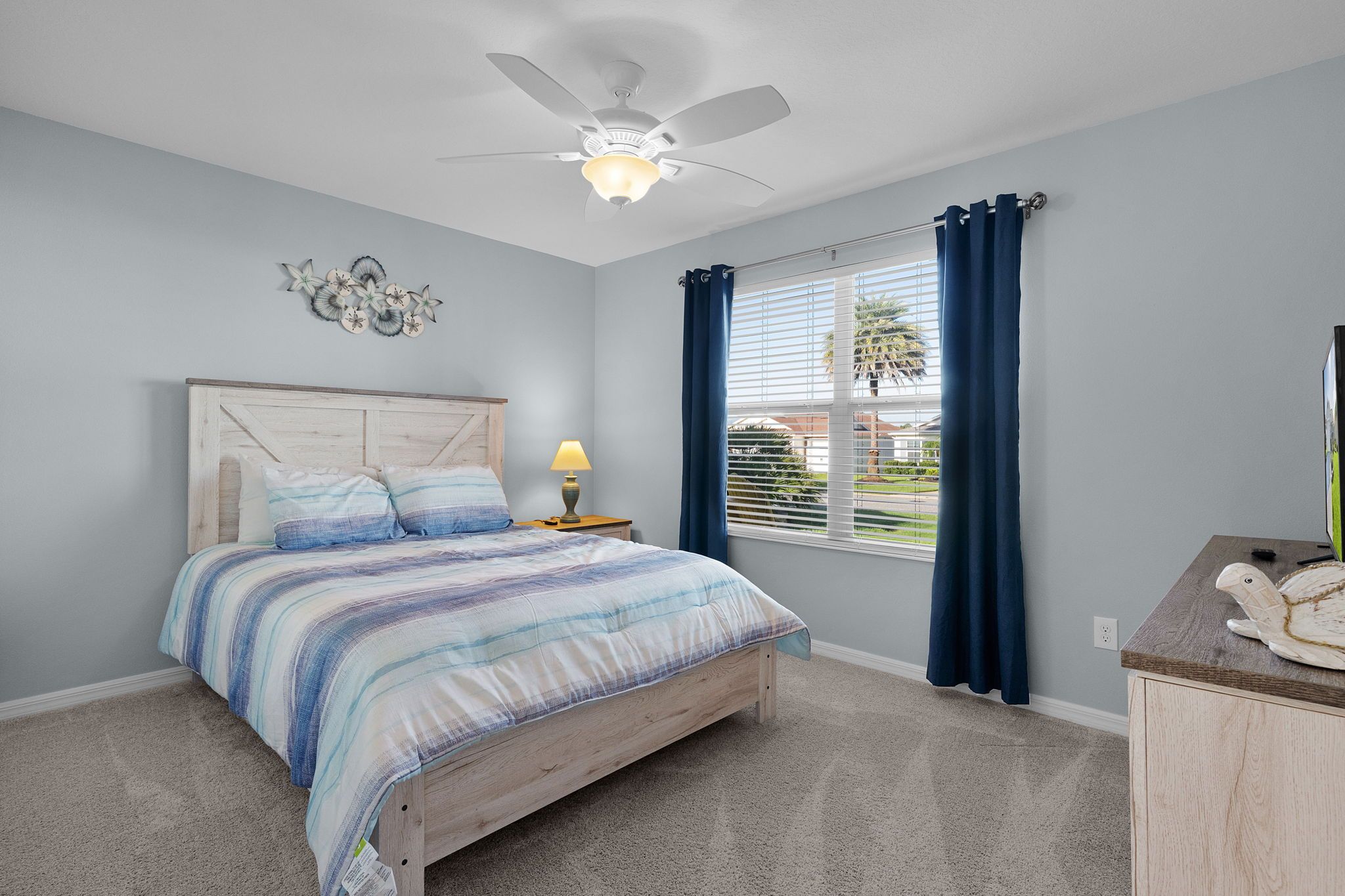 A bedroom with a bed, a ceiling fan, a window with curtains, and a nightstand with a lamp.
