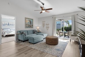 Ein modernes Wohnzimmer mit einer Eckcouch, Deckenventilator, großem Fenster und Blick ins Freie.