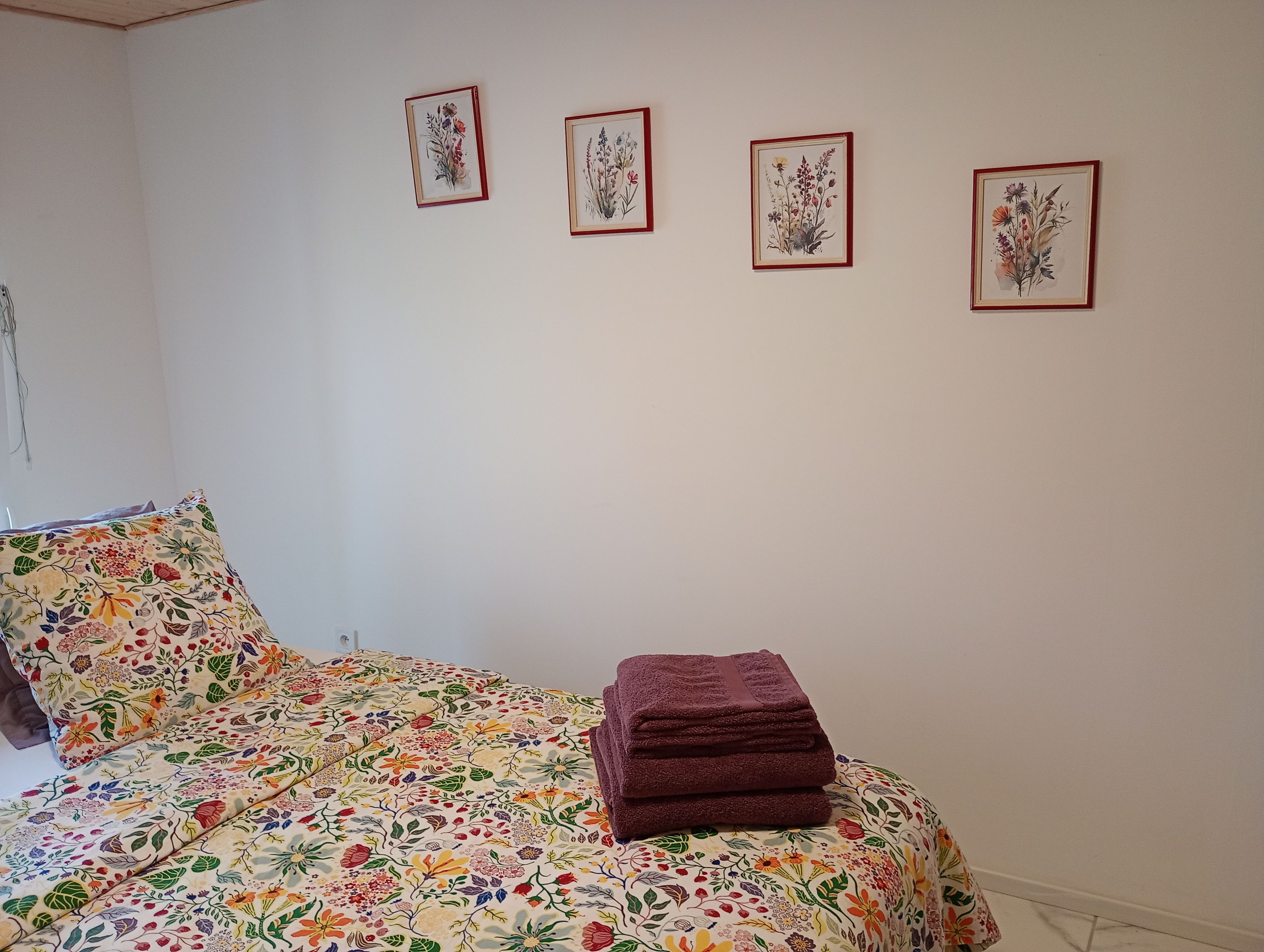 Un dormitorio con una colcha de estampado floral, una toalla oscura doblada con cuidado y cuatro láminas botánicas enmarcadas en la pared.