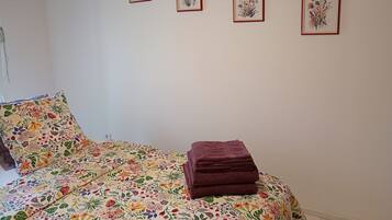 Un dormitorio con una colcha de estampado floral, una toalla oscura doblada con cuidado y cuatro láminas botánicas enmarcadas en la pared.