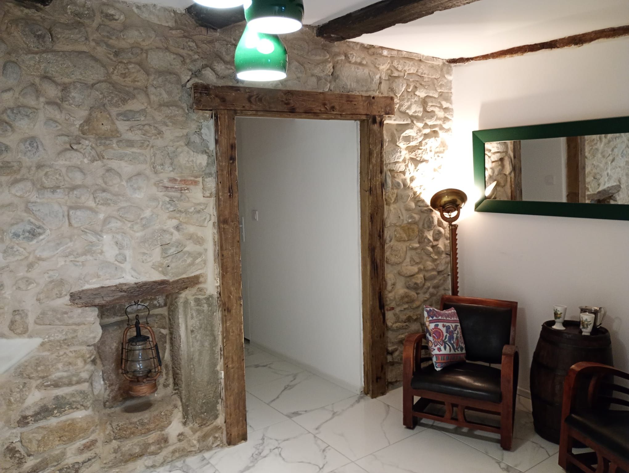 Habitació rústica amb parets de pedra, una porta de fusta, una làmpada penjant verda, un fanal tradicional, una cadira de pell i un barril de fusta.
