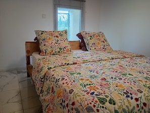 Un lit avec une couvre-lit à motifs floraux et deux oreillers, une tête de lit en bois, une fenêtre avec des stores et un sol carrelé.