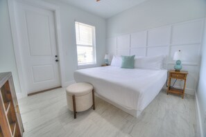 Ein Schlafzimmer mit einem großen Bett, einem kleinen runden Hocker, einem Nachttisch mit Lampe und einem Fenster mit Jalousien.
