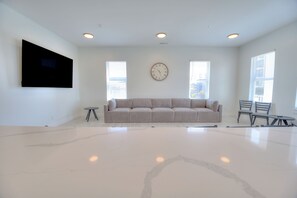 A spacious living room with a large flat-screen TV mounted on the wall, a beige sofa, a small round table, and a dining area with chairs. - BRAND NEW ACROSS FROM OCEAN 12 beds/10 baths • Pvt Pool and Spa • Sleeps 28 (North Myrtle Beach)