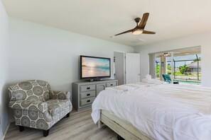 A bedroom with a bed, a television, a patterned armchair, a ceiling fan, and a view of the outdoors. - Turtle Cove | Heated Pool & Waterfront Retreat (Cape Coral)