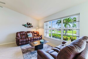 Sala de estar com sofá de couro marrom, mesa de centro e uma janela grande com vista para um jardim.