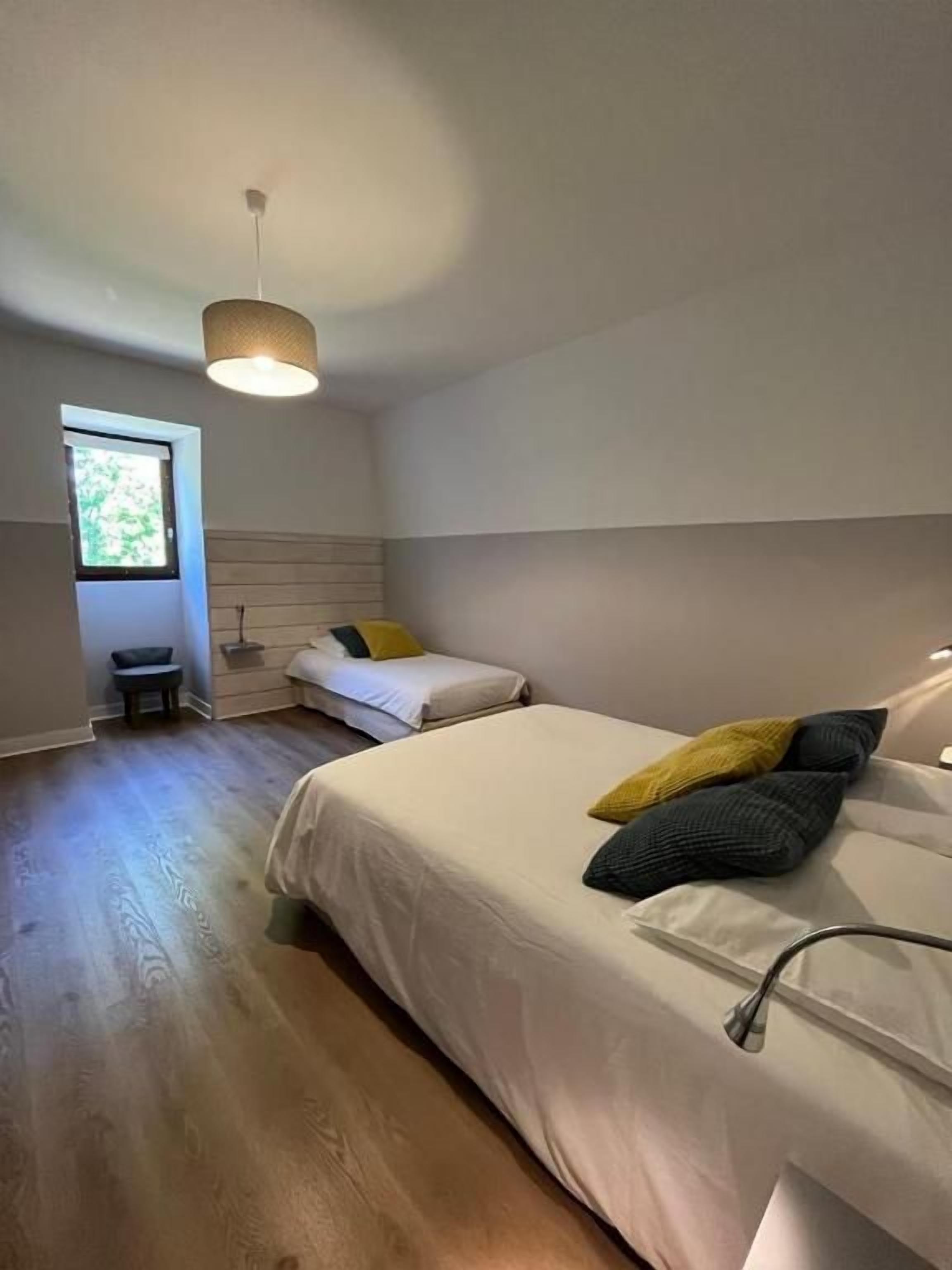 A bedroom with two beds, a bedside table, a lamp, a window, and a hanging light fixture.