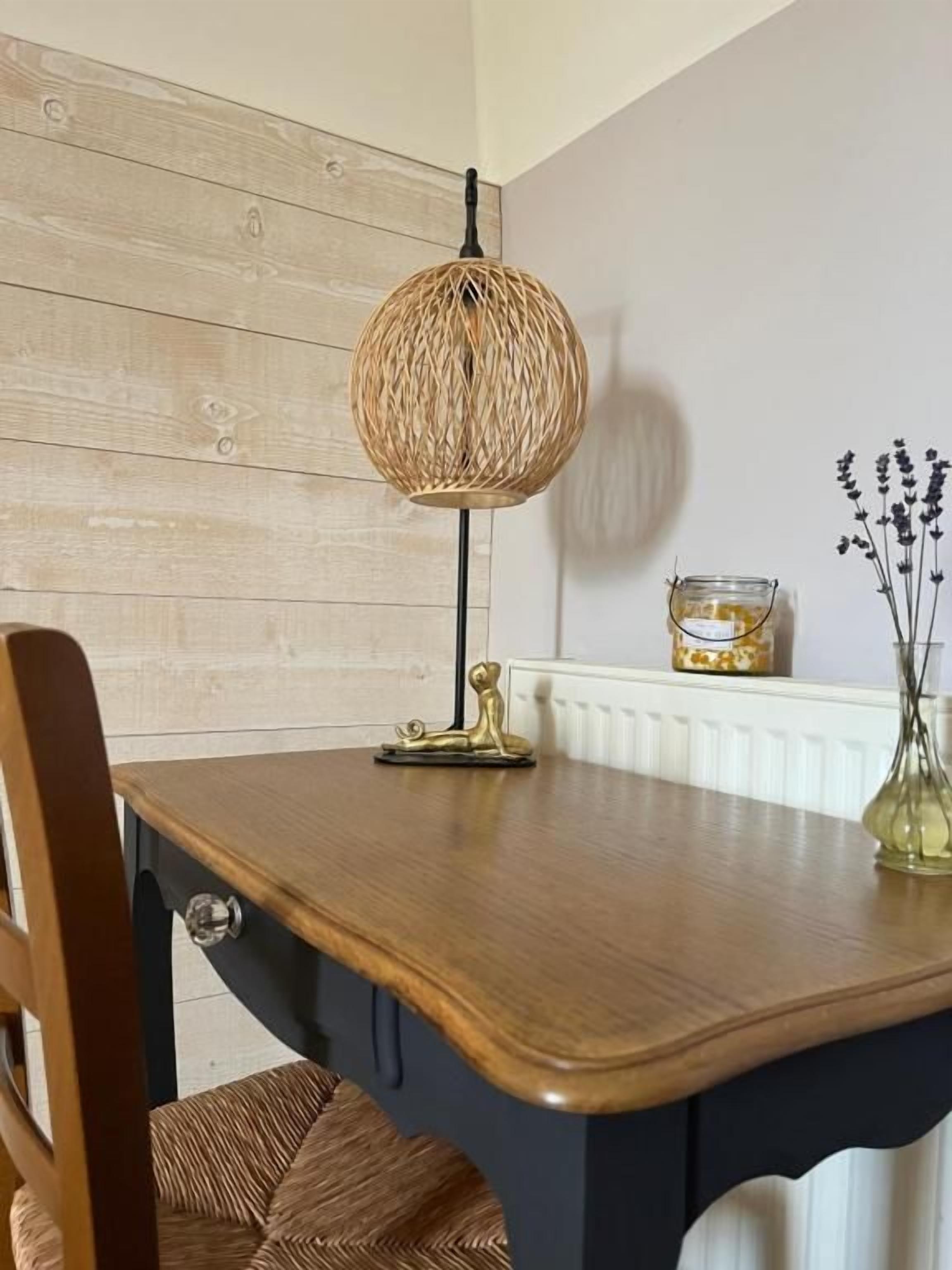 Une table à manger agrémentée d’un luminaire suspendu en fibres tressées, d’un vase contenant des fleurs séchées et d’un petit objet décoratif posé sur la table.