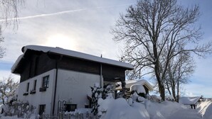 A two-story building with a sloped roof covered in snow, surrounded by snowbanks and leafless trees. - Grieshaber Haisl (Bad Bayersoien)