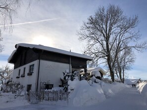 A two-story building with a sloped roof covered in snow, surrounded by snowbanks and leafless trees. - Grieshaber Haisl (Bad Bayersoien)