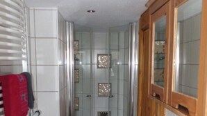 A narrow bathroom with a shower, a sink, and a wooden cabinet. - Grieshaber Haisl (Bad Bayersoien)
