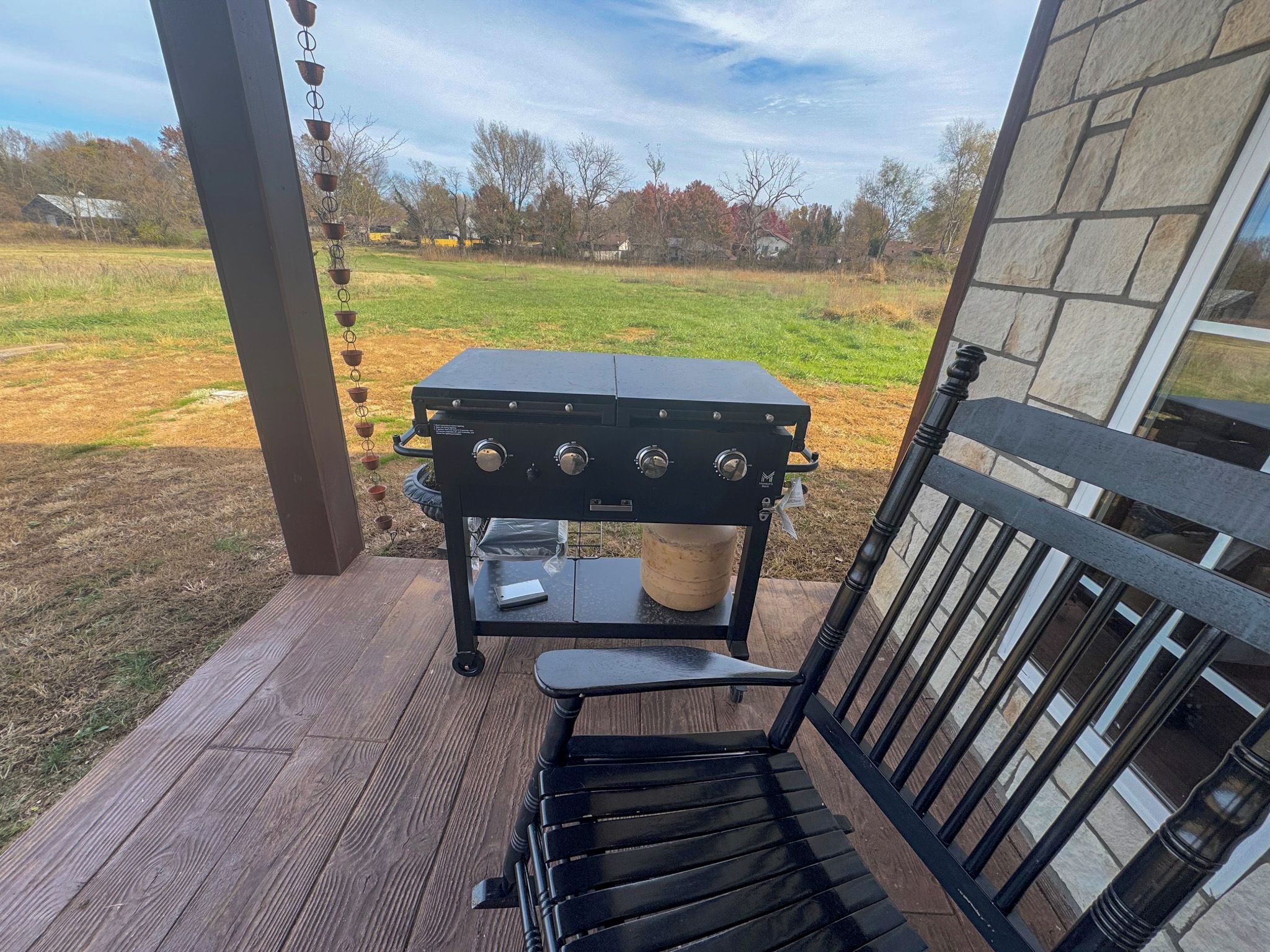 A barbecue grill on a wooden deck with a view of a grassy field and trees.
