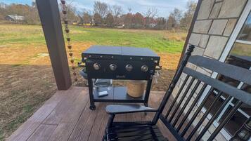 A barbecue grill on a wooden deck with a view of a grassy field and trees.
