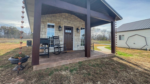 A covered patio with a stone facade, a wooden door, and a seating area with a table and chairs. - Quaint Cabin on 30 acres in Springfield (Springfield)