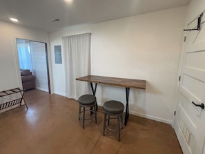 Quarto com mesa de madeira, dois banquinhos, porta branca e janela com cortinas.