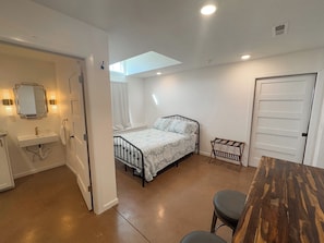 Quarto com cama, uma mesinha, um banco, um espelho e uma porta.