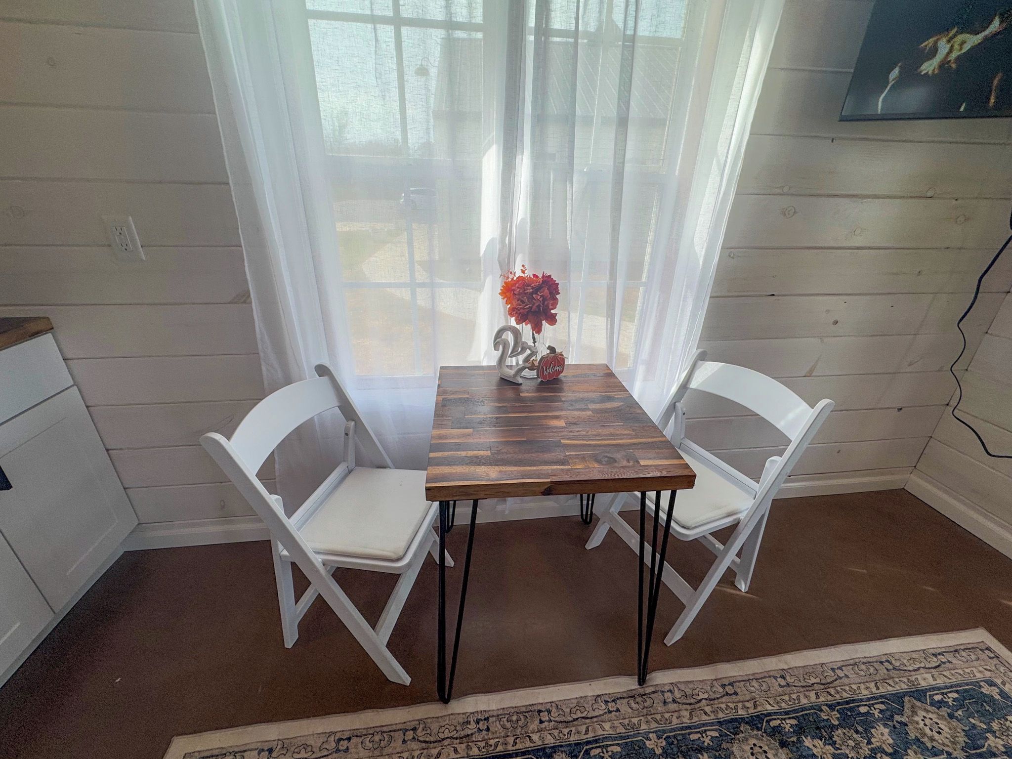 Een eethoek met een houten tafel en twee witte stoelen, een vaas met bloemen en uitzicht door dunne gordijnen.