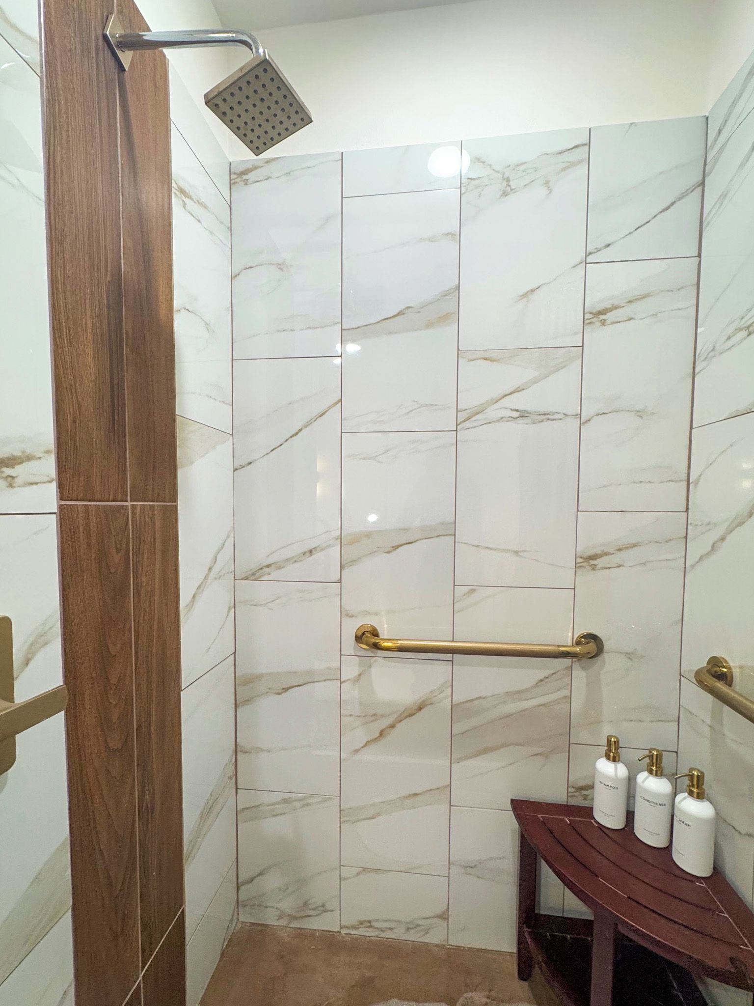 A shower area with a marble wall, a wooden door frame, a showerhead, and a handrail.