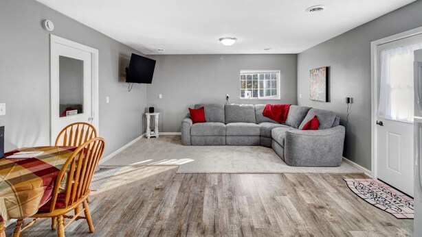 Een woonkamer met een grijze hoekbank, een houten eettafel met stoelen, een flatscreen televisie aan de muur en een raam met witte gordijnen.