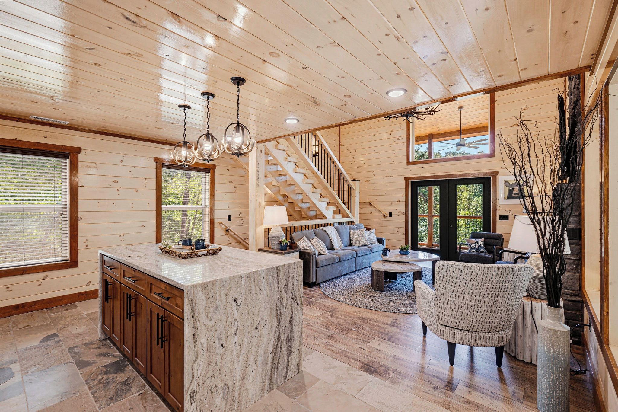 A spacious living area with a wooden interior, a marble island, a staircase, and a view of the outdoors.