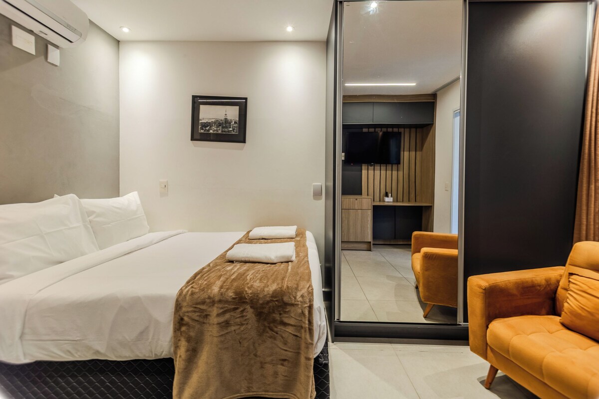 Quarto de hotel com cama, sofá, televisão e banheiro visível por uma porta de vidro.