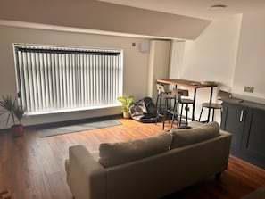 Una sala moderna con sofá, mesa de comedor con sillas, planta en maceta y ventana con persianas.