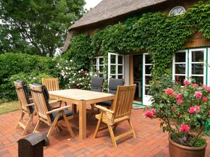 Eine Terrasse mit Holztisch und -stühlen, umgeben von einem Backsteinboden, und ein Haus mit einem Reetdach und grünem Efeu.