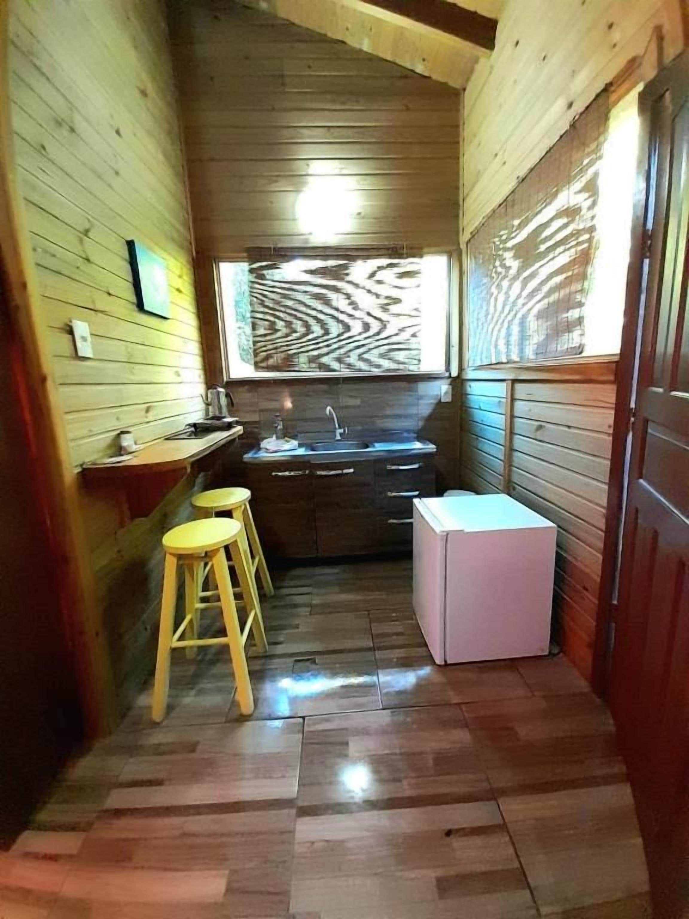 A narrow wooden room with a sink, stools, and a refrigerator.