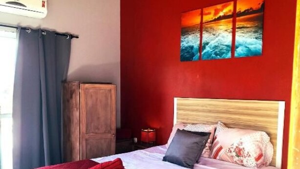 A bed with a red blanket, a wooden headboard, and a painting of a sunset on the wall. - Pousada Baia Rosa (Imbituba)