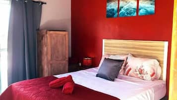 Un lit avec une couverture rouge, une tête de lit en bois et un tableau représentant un coucher de soleil accroché au mur.