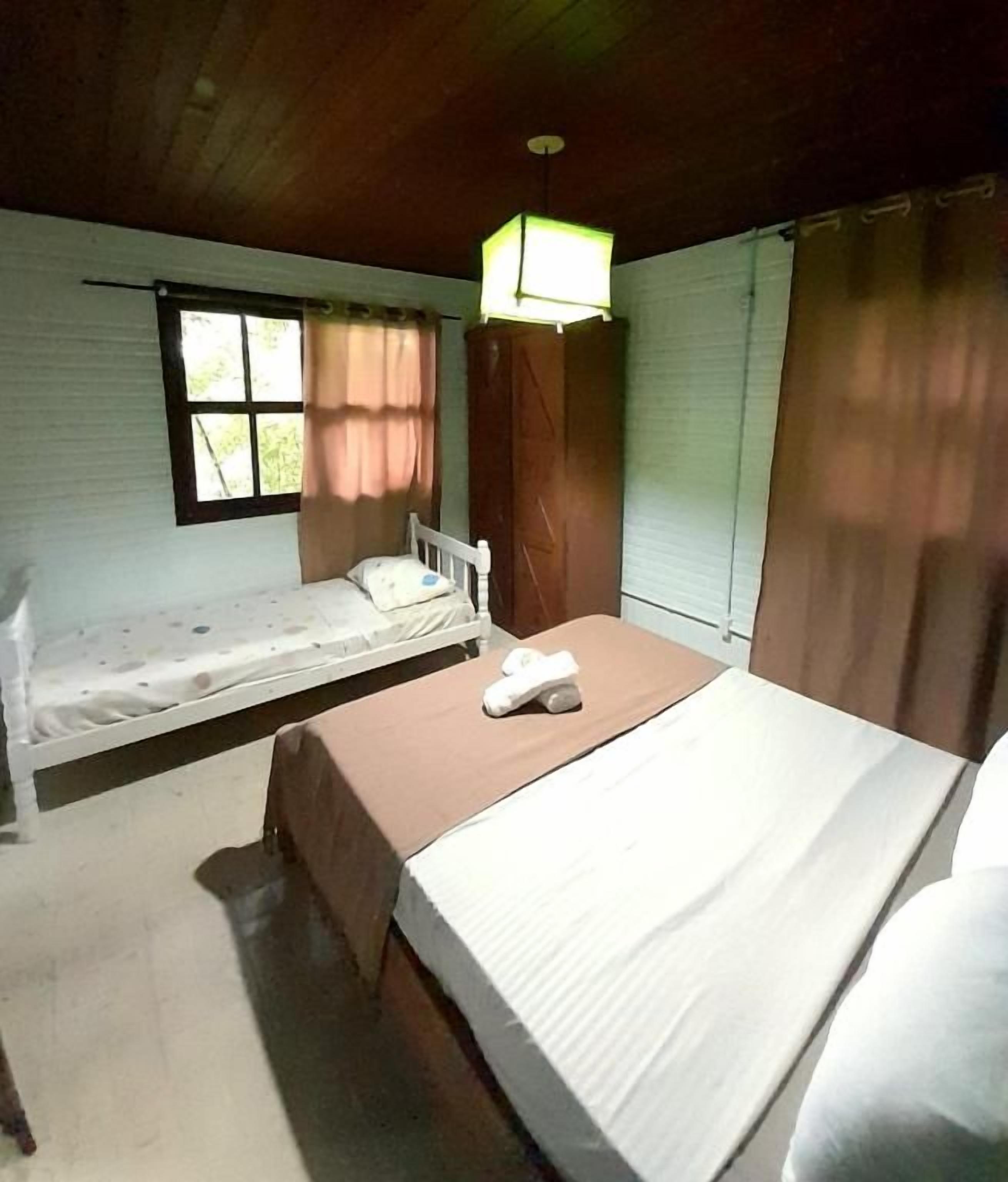 Una habitación con dos camas, una con cabecera blanca y la otra con cabecera de madera. Hay una lámpara sobre las camas y una ventana con cortinas a la izquierda.
