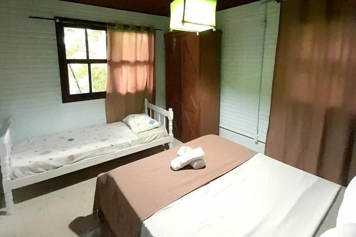 Ein Zimmer mit zwei Betten, eines mit einem weißen Kopfteil und das andere mit einem Holzkopfteil. Über den Betten hängt eine einzelne Leuchte, und links befindet sich ein Fenster mit Vorhängen.
