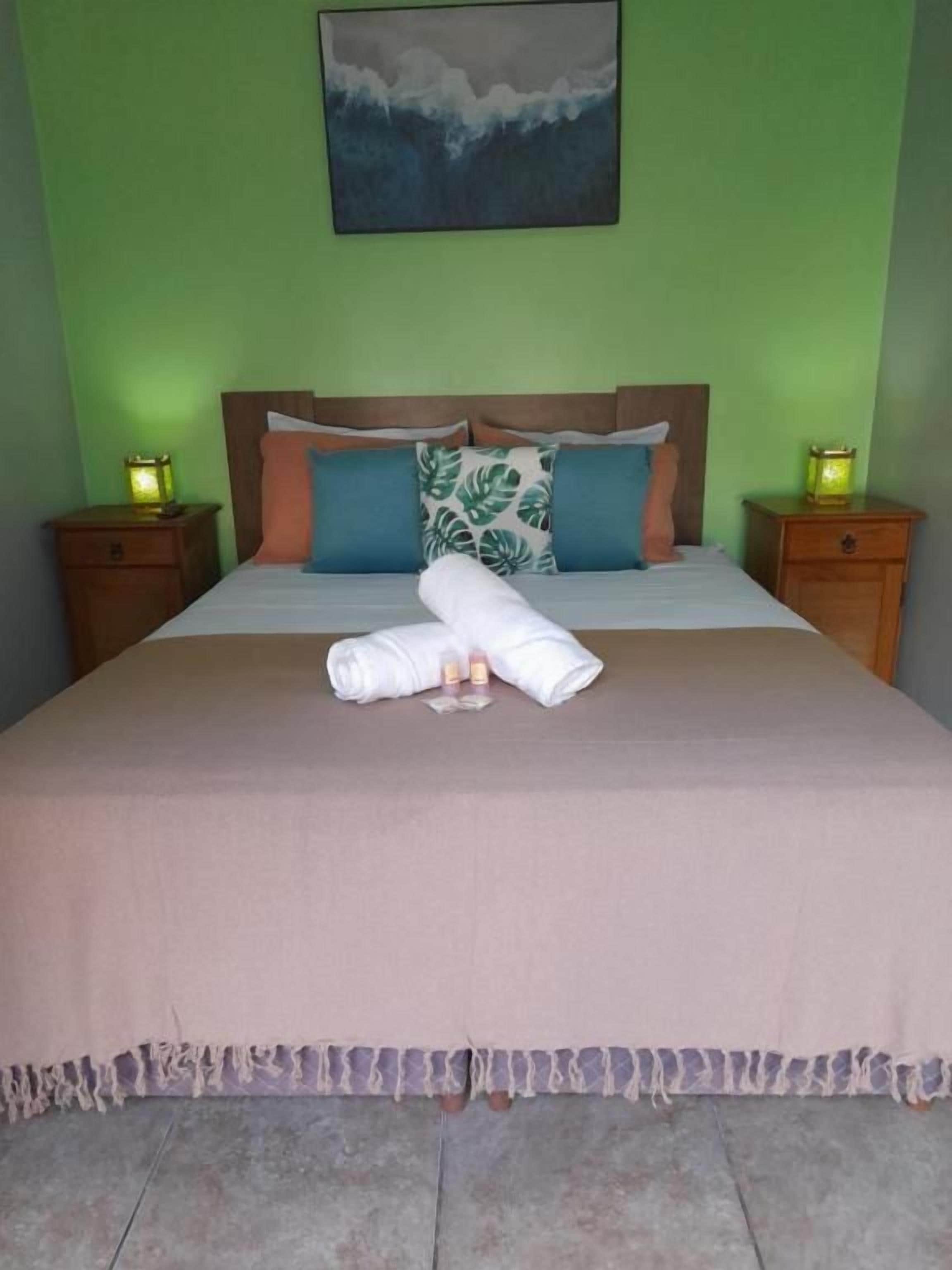 Una cama bien hecha con una colcha rosa y una toalla blanca encima. Hay dos mesitas de noche con lámparas, y un cuadro cuelga sobre la cama.