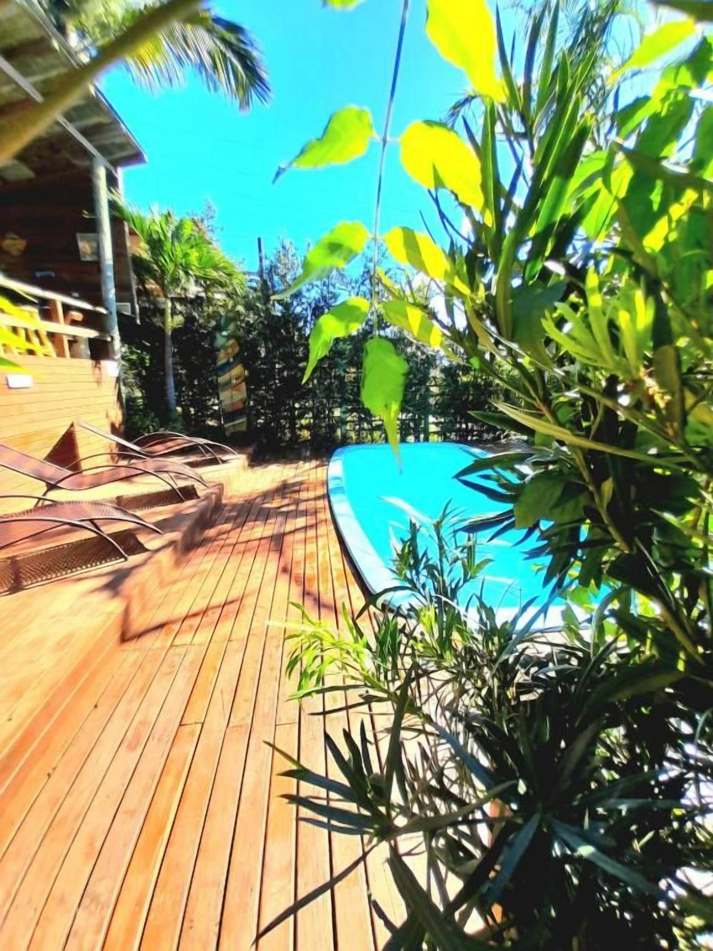 A wooden deck with a pool, surrounded by greenery and a building in the background.