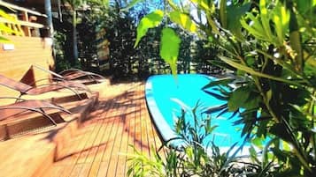 A wooden deck with a pool, surrounded by greenery and a building in the background.