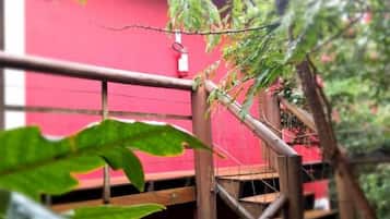 Una escalera de madera con barandilla de metal, rodeada de follaje y una pared roja.