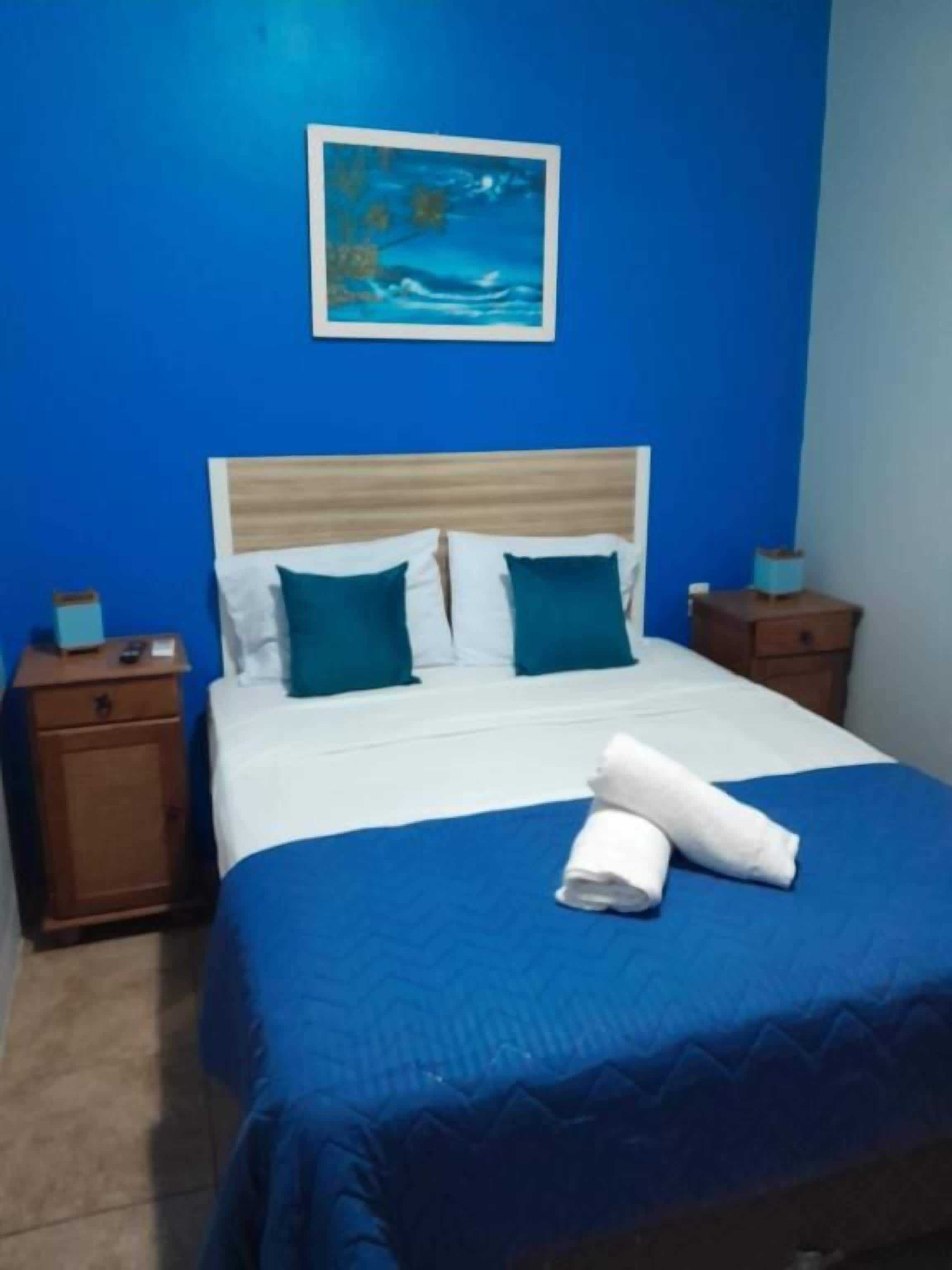 Habitación de hotel con cama, paredes azules, un cuadro y una mesita de noche con lámpara.