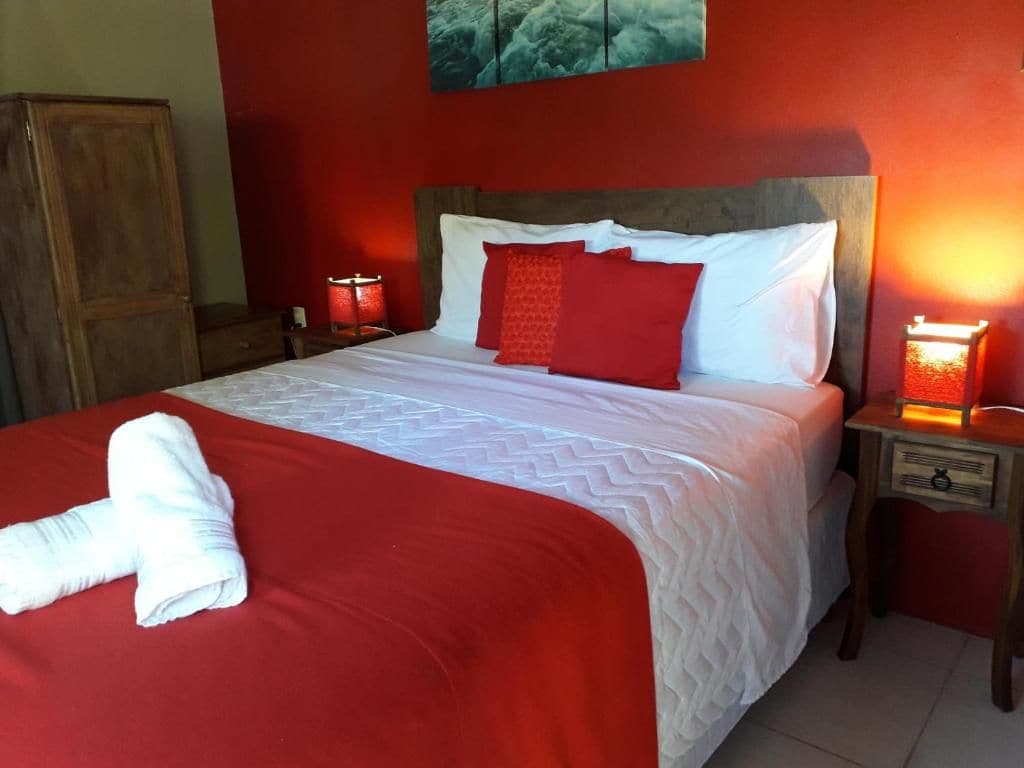 Una cama con una colcha roja y blanca, dos almohadas rojas y una toalla blanca sobre la cama. Hay dos lámparas de noche a cada lado de la cama.