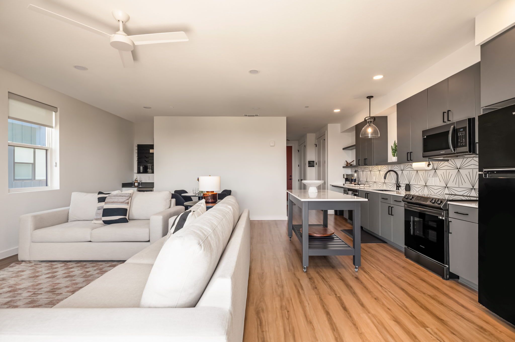 A modern kitchen with a black refrigerator, microwave, and oven. There is a dining table with a lamp and a sofa in the living area.