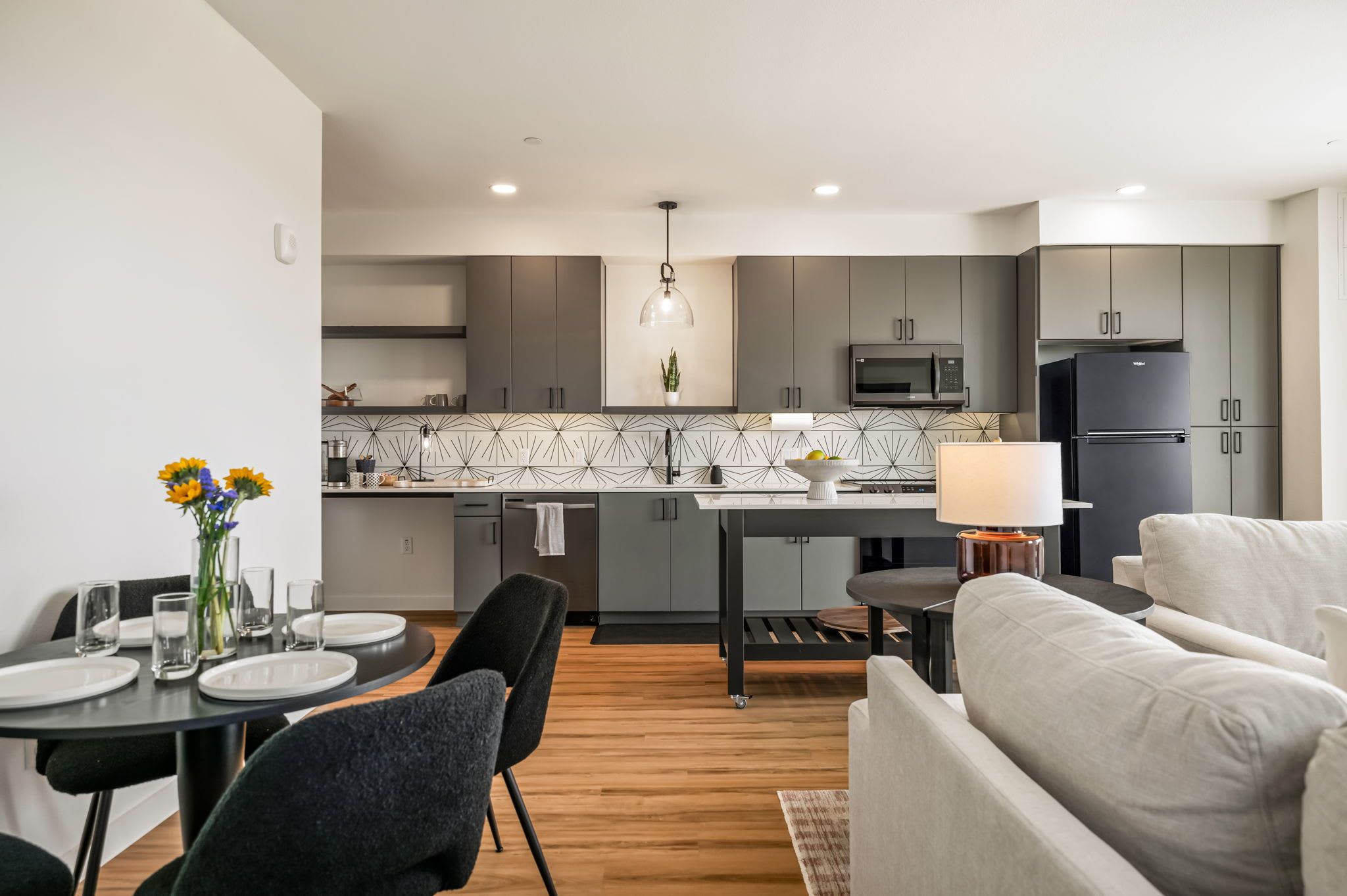 A modern kitchen with grey cabinets, a microwave, and a refrigerator. There is a dining area with a table set for two, a sofa, and a lamp.