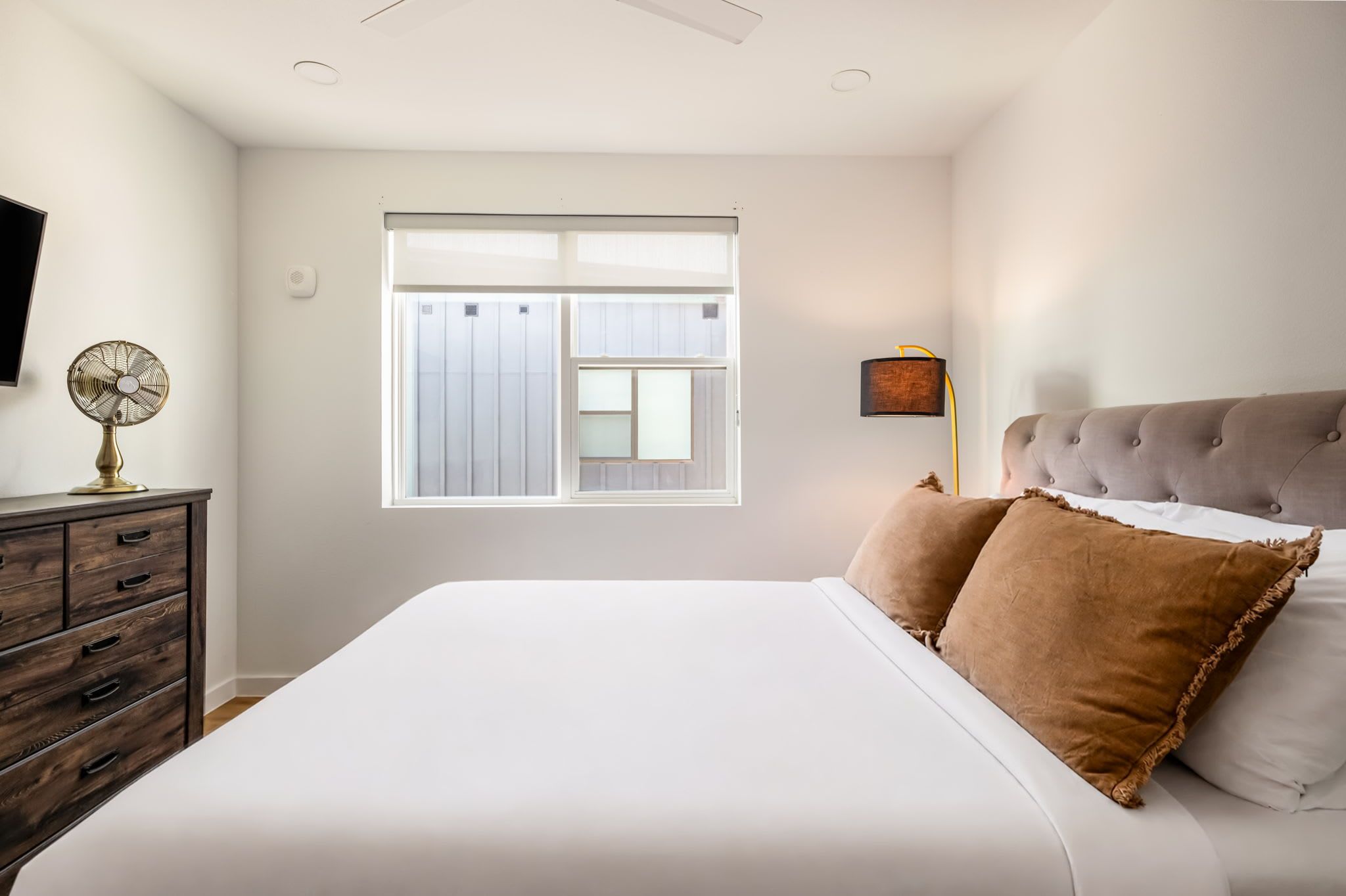 A bedroom with a large bed, a dresser, a window with blinds, and a wall-mounted lamp.