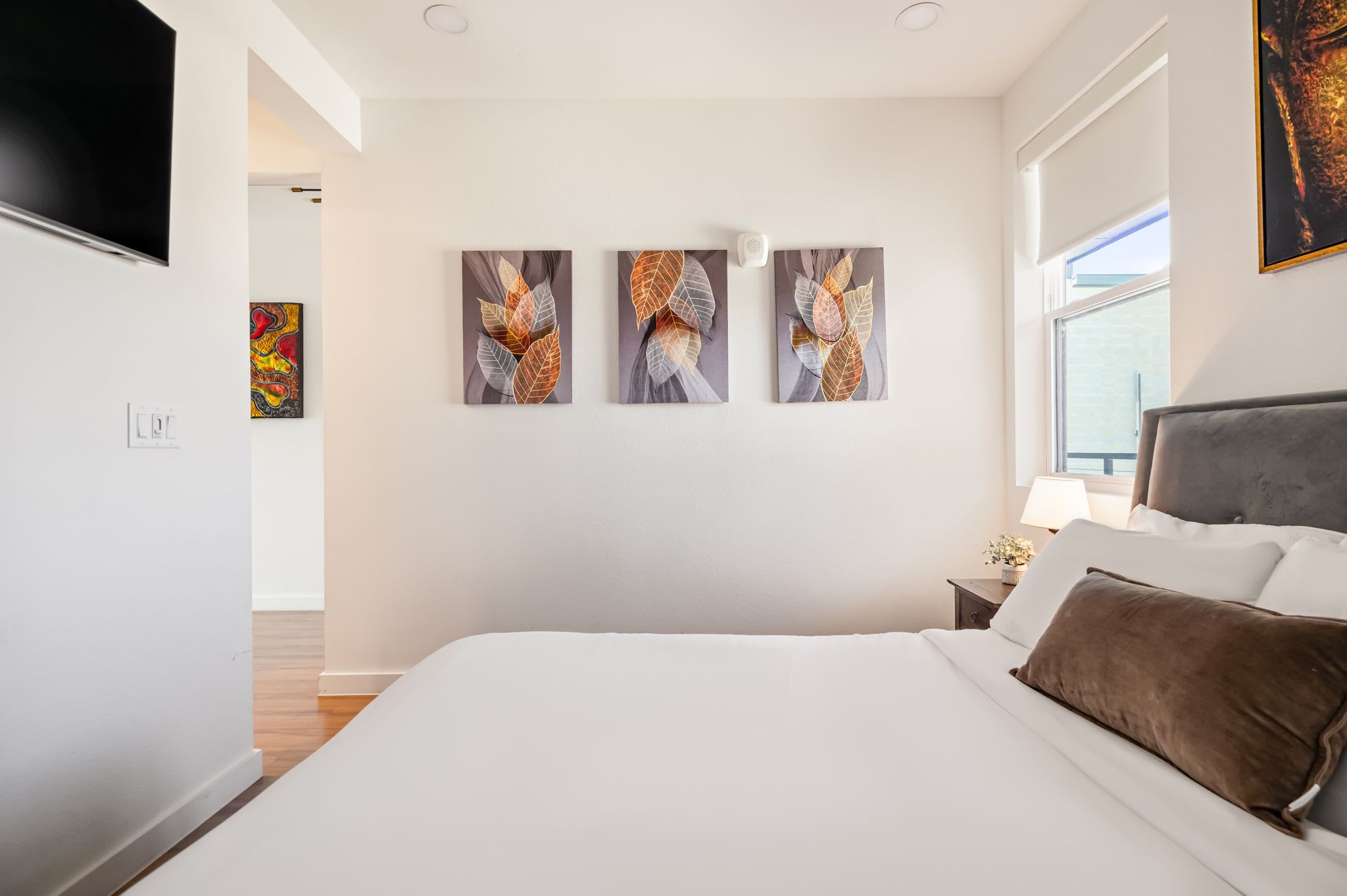 A modern bedroom with a large bed, a nightstand, a lamp, and three wall-mounted paintings.