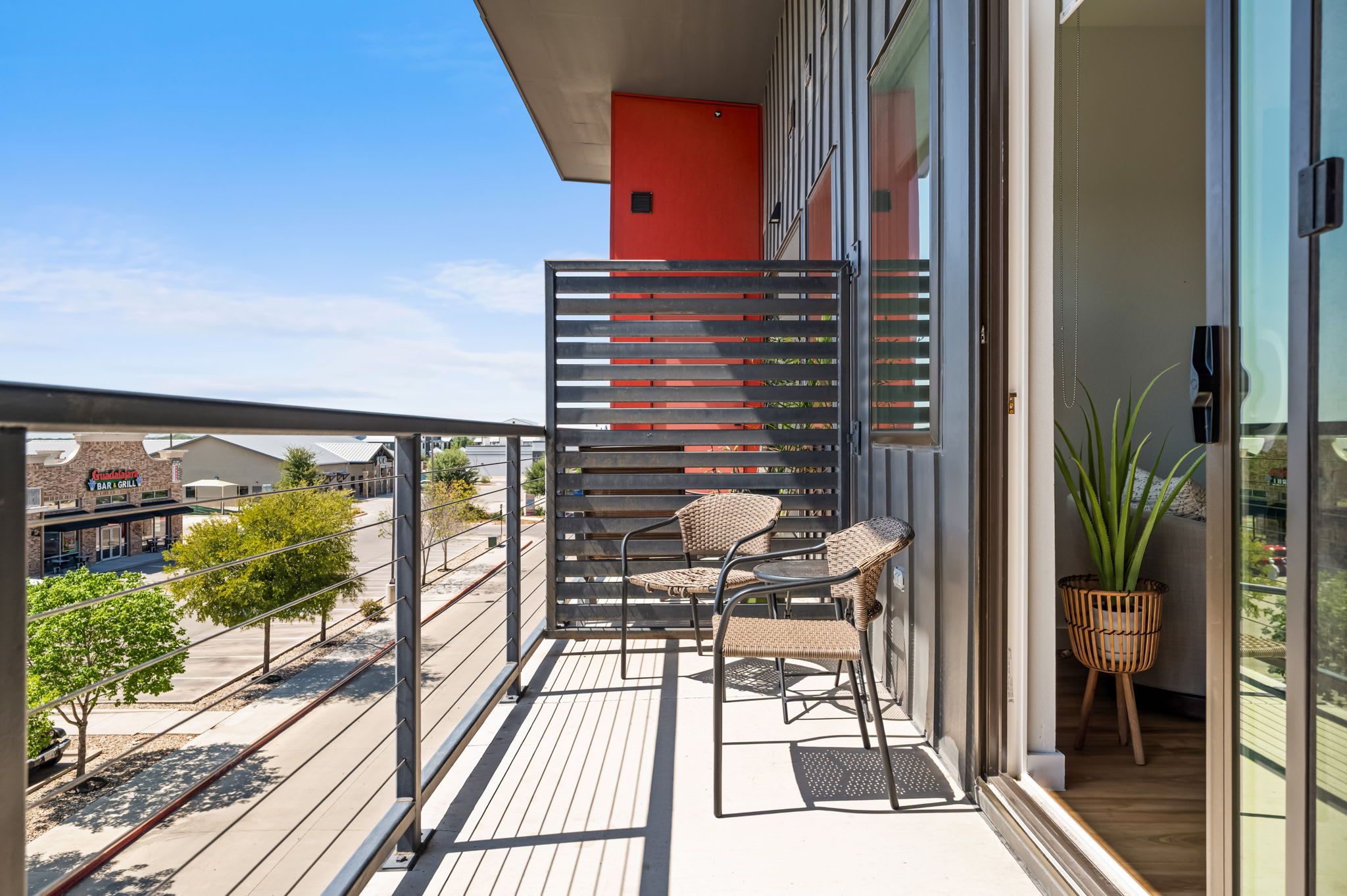A balcony with a metal railing, two wicker chairs, a small table, and a potted plant.