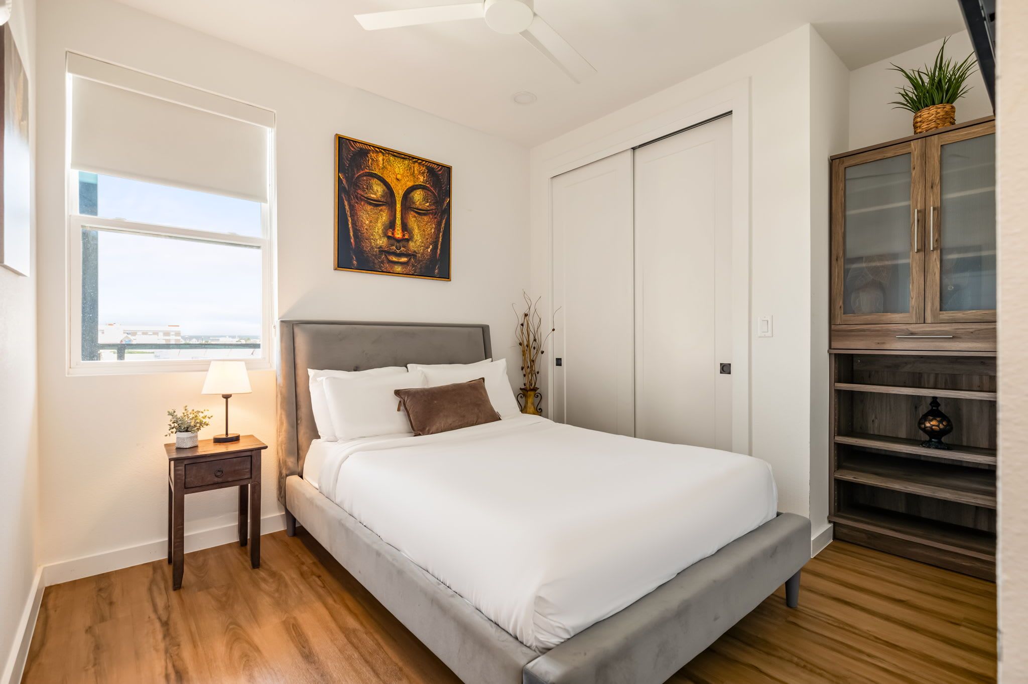 Een slaapkamer met een bed, nachtkastje, lamp, kunstwerk, garderobe en uitzicht op de stad.