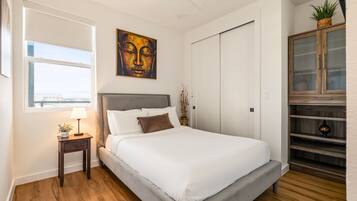Een slaapkamer met een bed, nachtkastje, lamp, kunstwerk, garderobe en uitzicht op de stad.