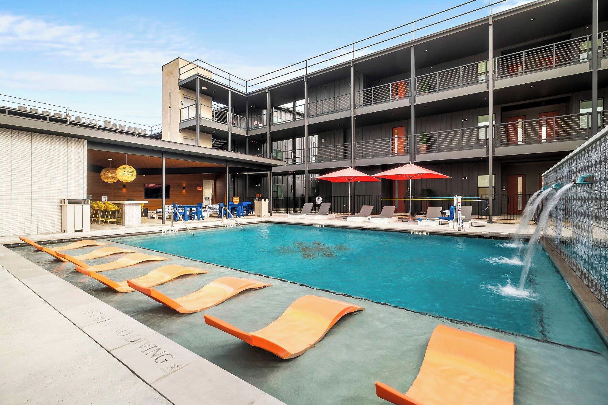 A modern pool area with lounge chairs and umbrellas, a bar area, and a multi-story building with balconies.
